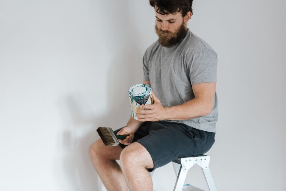 Crop young bearded male in casual wear with paint brush sitting near white wall in house